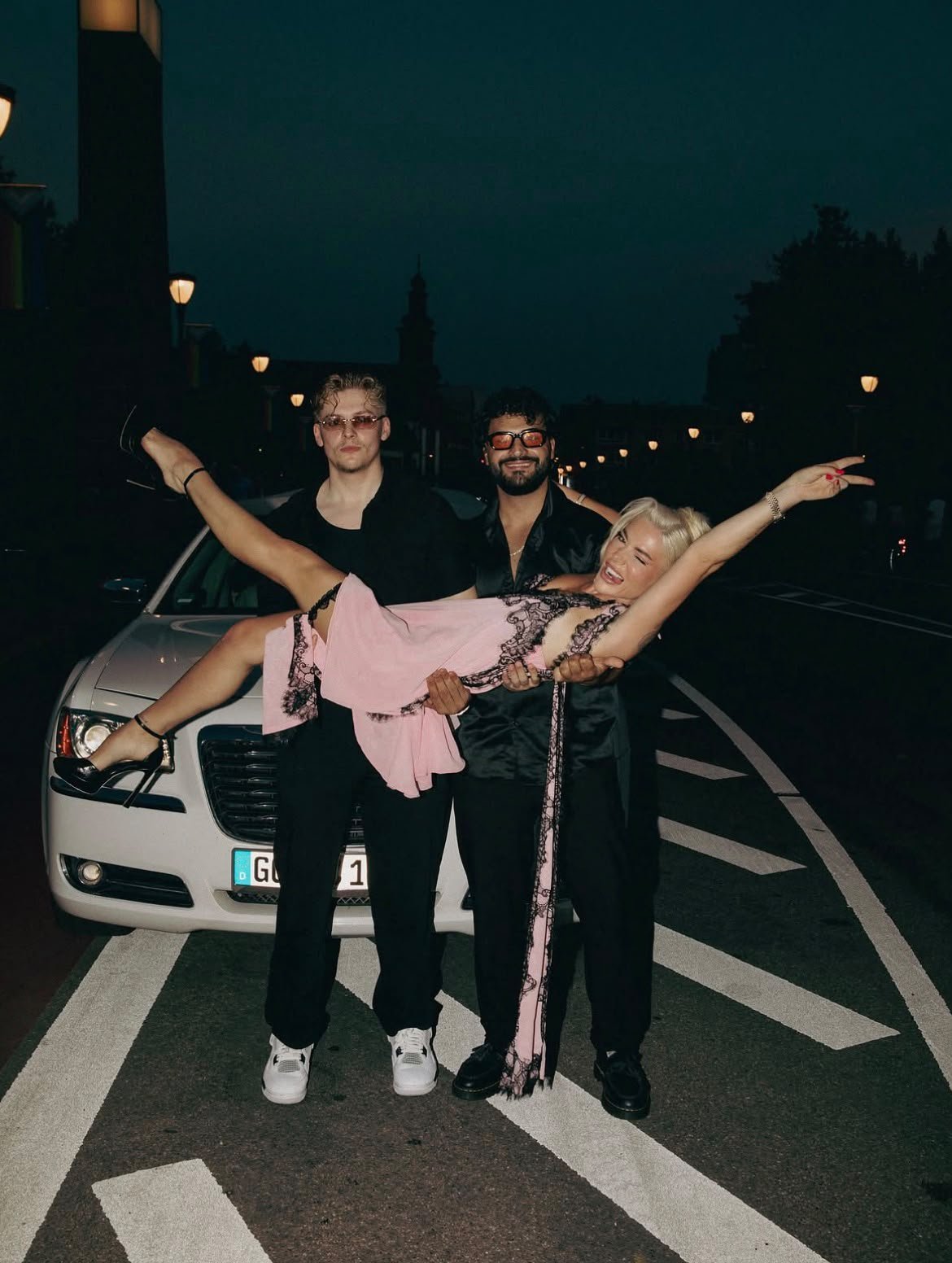 Three people pose playfully in front of a white car on a nighttime city street with illuminated lamp posts in the background